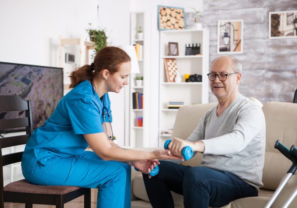 Female nurse doing physiotherapy with senior man in nursing home using dumbbells.